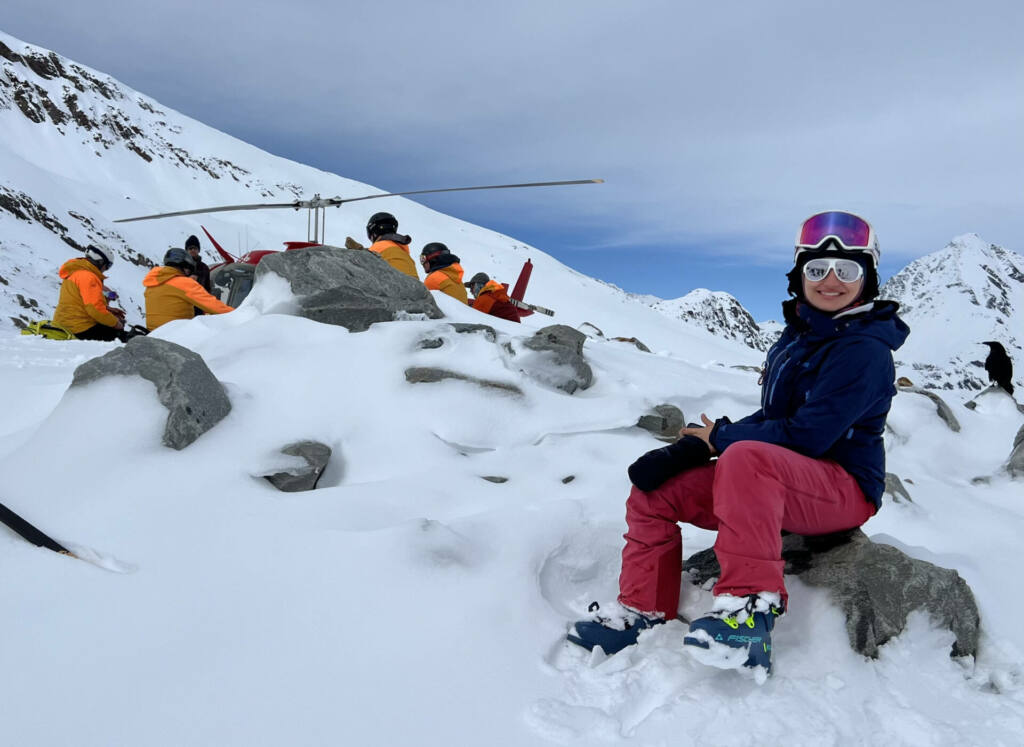 Mulher sorridente sentada sobre uma rocha coberta de neve em uma paisagem montanhosa. Ela usa roupas de ski — calça rosa, jaqueta azul e botas de neve — e óculos espelhados. Ao fundo, um grupo de pessoas com jaquetas amarelas se reúne ao redor de um helicóptero vermelho em meio às montanhas cobertas de neve.